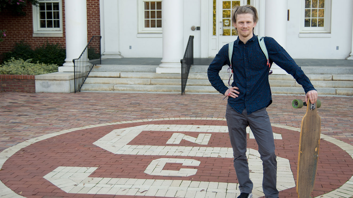 Carter Pape standing with his longboard in front of Winslow Hall at NC State University