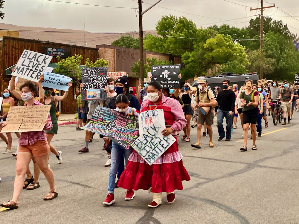 people including a woman dressed in traditional Native American attire walk down Center Street in Moab, Utah carrying signs calling for racial justice
