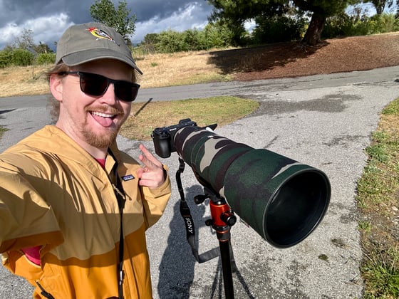 Carter throws up a peace sign next to a camera and telephoto lens, which is wrapped in camouflage